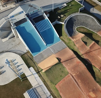 An aerial view of an outdoor sports facility featuring a blue skateboard ramp, a winding BMX track with dirt surfaces, and adjacent walking areas with seating and pathways. The surrounding area shows green grass and paved sections, with shadows indicating the presence of tall structures or lighting poles.
