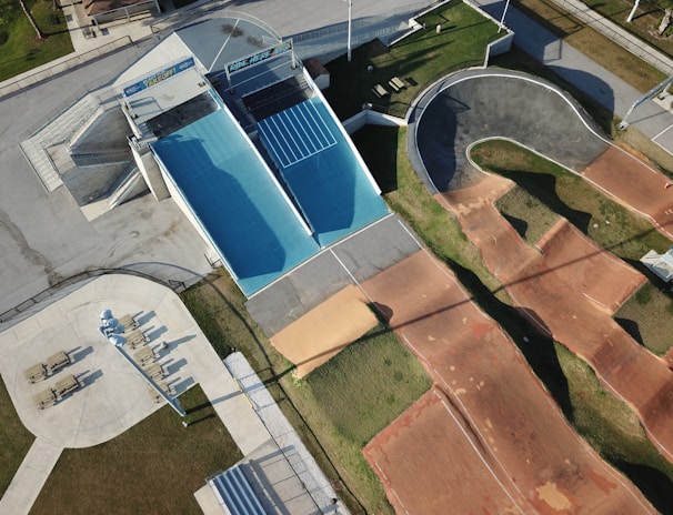 An aerial view of an outdoor sports facility featuring a blue skateboard ramp, a winding BMX track with dirt surfaces, and adjacent walking areas with seating and pathways. The surrounding area shows green grass and paved sections, with shadows indicating the presence of tall structures or lighting poles.