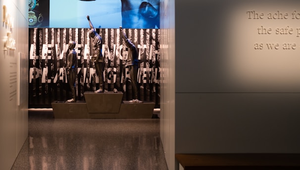 A dimly lit display features statues of athletes standing on a podium. They are positioned in front of a backdrop with the words 'LEVELING THE PLAYING FIELD' in bold letters. On the wall to the right, a softly lit quote is partially visible. The overall ambiance suggests a museum or exhibition setting.