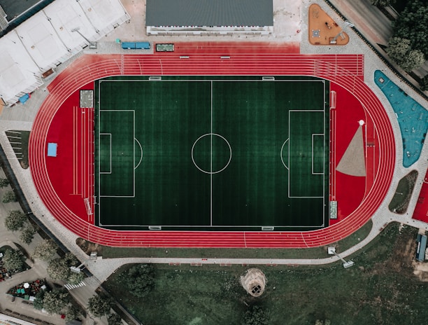 An aerial view of a large sports complex featuring a central green football field surrounded by a red running track. Adjacent to the track, there is a parking area, buildings, and pathways. Nearby, there are additional sports courts in different colors, including a blue swimming pool and orange basketball courts.