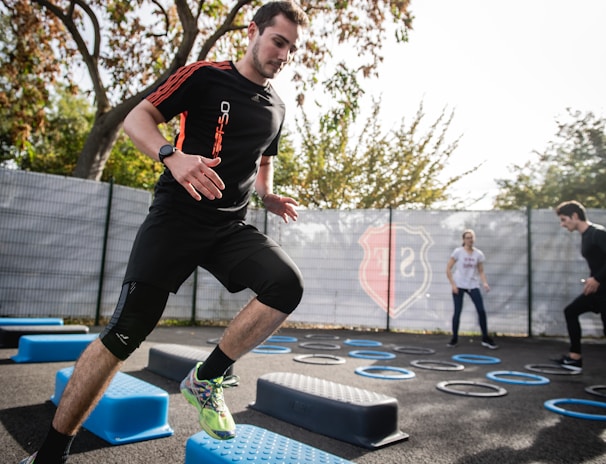 A man is actively participating in an outdoor fitness session, jumping over blue platforms with agility. Two other people in the background are also engaging in exercise, with rings scattered on the ground. The area is fenced and surrounded by trees, suggesting a park or recreational setting.