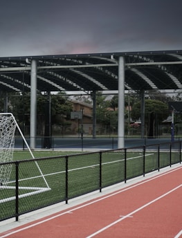 An outdoor sports facility featuring a running track and a soccer goal. The track is red, surrounded by a black metal fence, and the field is covered with artificial green turf. In the background, a basketball court is visible under a covered roof with metal supports. The sky is overcast, adding a moody atmosphere.