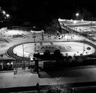 A nighttime aerial view of an outdoor recreational facility with various sports and play areas. The foreground features a running track with the words 'PARK G3' written on it. Surrounding the track are playground structures and a round seating area. Further in the background, several tennis courts and a basketball court are visible, with bright lights illuminating the entire area.