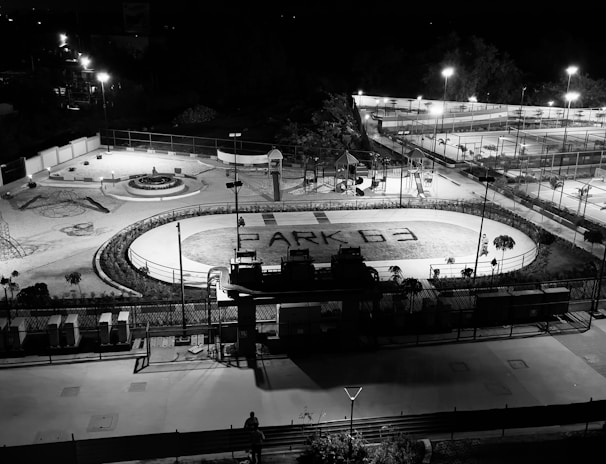 A nighttime aerial view of an outdoor recreational facility with various sports and play areas. The foreground features a running track with the words 'PARK G3' written on it. Surrounding the track are playground structures and a round seating area. Further in the background, several tennis courts and a basketball court are visible, with bright lights illuminating the entire area.