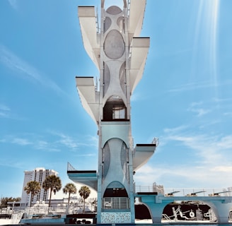 A tall, modern diving platform structure stands against a clear blue sky. The platform has multiple levels with geometric cutouts and sleek design elements. Below it is a large, inviting swimming pool with clear blue water. In the background, there are palm trees and a few multi-story buildings, adding an urban backdrop to the scene.
