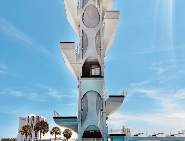 A tall, modern diving platform structure stands against a clear blue sky. The platform has multiple levels with geometric cutouts and sleek design elements. Below it is a large, inviting swimming pool with clear blue water. In the background, there are palm trees and a few multi-story buildings, adding an urban backdrop to the scene.