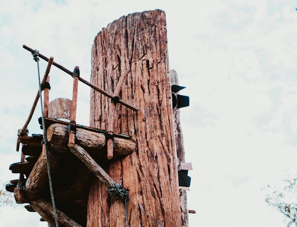A tall, weathered wooden post supports a rustic platform constructed from wooden planks and ropes. The structure appears part of an adventure or obstacle course, with ladders or steps attached to the post. The sky is partly cloudy, and some tree branches can be seen in the background.