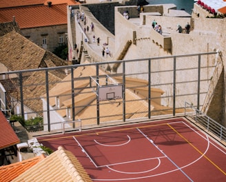 A vibrant red sports court is situated on a rooftop, surrounded by terracotta-tiled buildings overlooking a historic stone wall that stretches alongside a beautiful coastal view. People walk along the stone path near the sea, suggesting a popular tourist spot or cultural landmark. The setting combines elements of modern recreation with historic architecture, creating a unique juxtaposition.