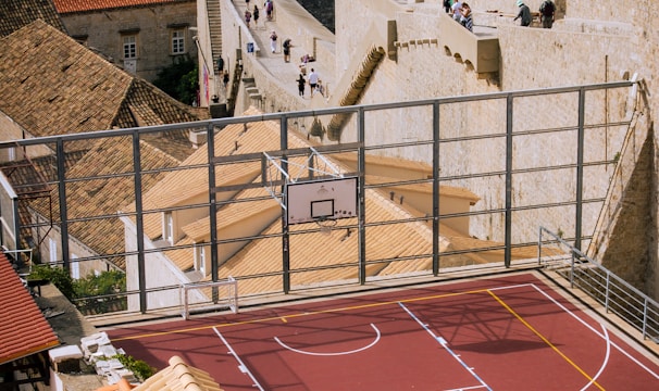 A vibrant red sports court is situated on a rooftop, surrounded by terracotta-tiled buildings overlooking a historic stone wall that stretches alongside a beautiful coastal view. People walk along the stone path near the sea, suggesting a popular tourist spot or cultural landmark. The setting combines elements of modern recreation with historic architecture, creating a unique juxtaposition.