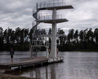A tall diving platform stands beside a body of water. The platform has three levels with a spiral staircase on one side. There are three people near the platform, two standing on the dock and one walking away. The scene is set under a cloudy sky with a row of trees visible in the background.