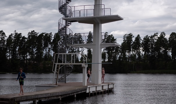 A tall diving platform stands beside a body of water. The platform has three levels with a spiral staircase on one side. There are three people near the platform, two standing on the dock and one walking away. The scene is set under a cloudy sky with a row of trees visible in the background.